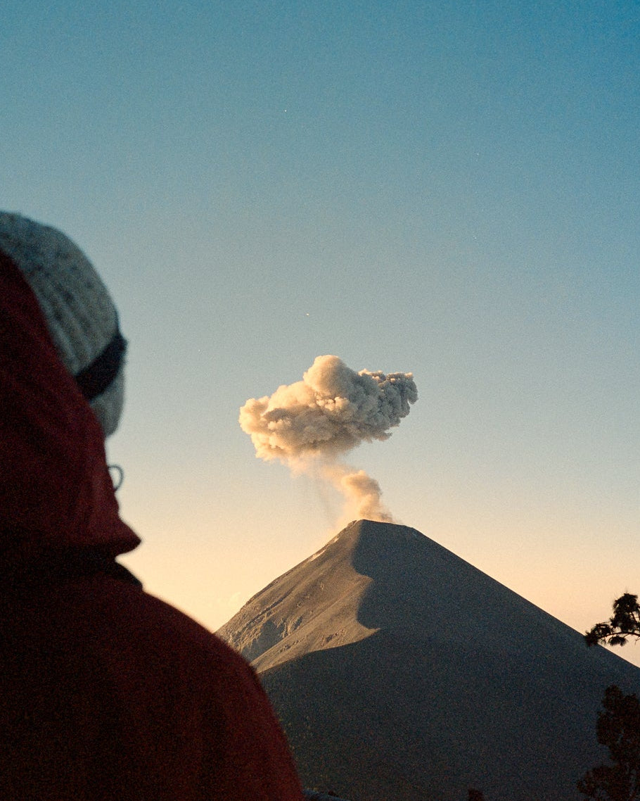A person in a red hooded jacket and white knit beanie looks toward a volcano erupting under a clear blue sky, with dark tree silhouettes at the right edge.