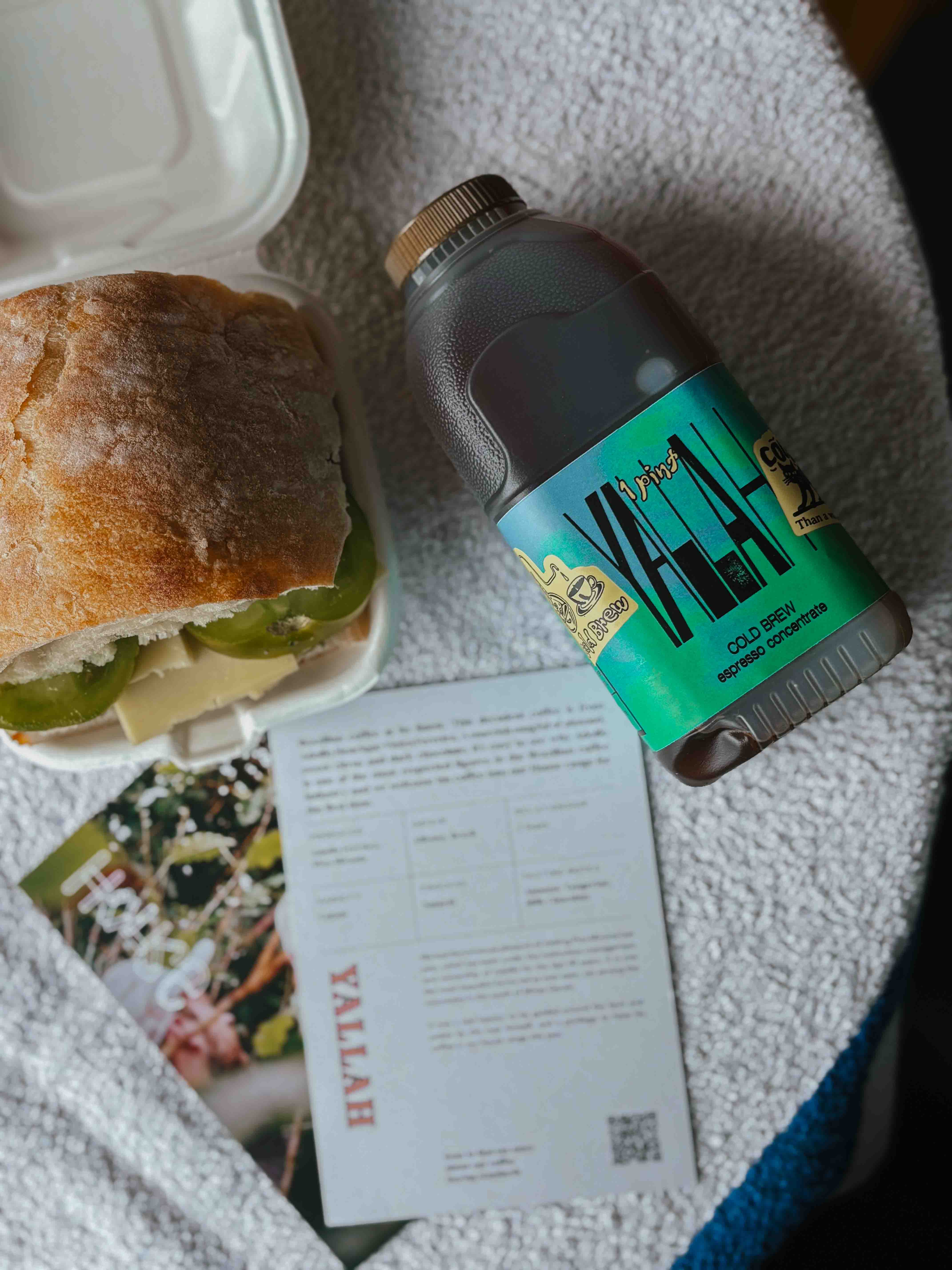 A bottle of Yallah Cold Brew 1 Pint coffee to a sandwich in a white container and a printed recipe card, arranged on a textured white towel.